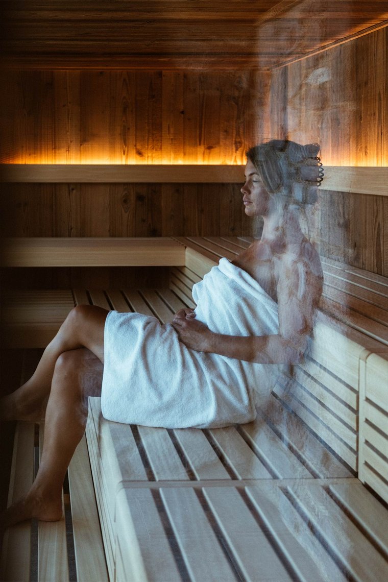 Eine Frau sitzt entspannt in einer Sauna, eingehüllt in ein weißes Handtuch. Sie hat die Augen geschlossen und genießt die Wärme. Der Holzraum ist sanft beleuchtet, was eine beruhigende Atmosphäre schafft. Dampfschwaden sind sichtbar, die die Stimmung unterstreichen.