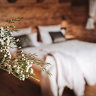 Ein gemütliches Schlafzimmer mit einem großen Bett, das mit weißen Bettwäsche und Kissen dekoriert ist. Im Vordergrund ist ein kleiner Zweig zu sehen, der das Bild ergänzt. Der Raum hat einen warmen, natürlichen Holzlook.