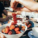 Eine Hand hält eine Traube über einer Schüssel mit frischen Beeren, darunter Erdbeeren, Himbeeren und Heidelbeeren. Im Hintergrund sind weitere Speisen und Getränke auf einem Tisch zu erkennen.