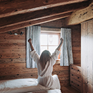 Eine Person sitzt auf einem Bett in einem rustikalen Zimmer mit Holzverkleidung. Sie hat die Arme in die Luft erhoben, als würde sie den Morgen begrüßen. Sonnenlicht strömt durch das Fenster, das mit Vorhängen geschmückt ist.
