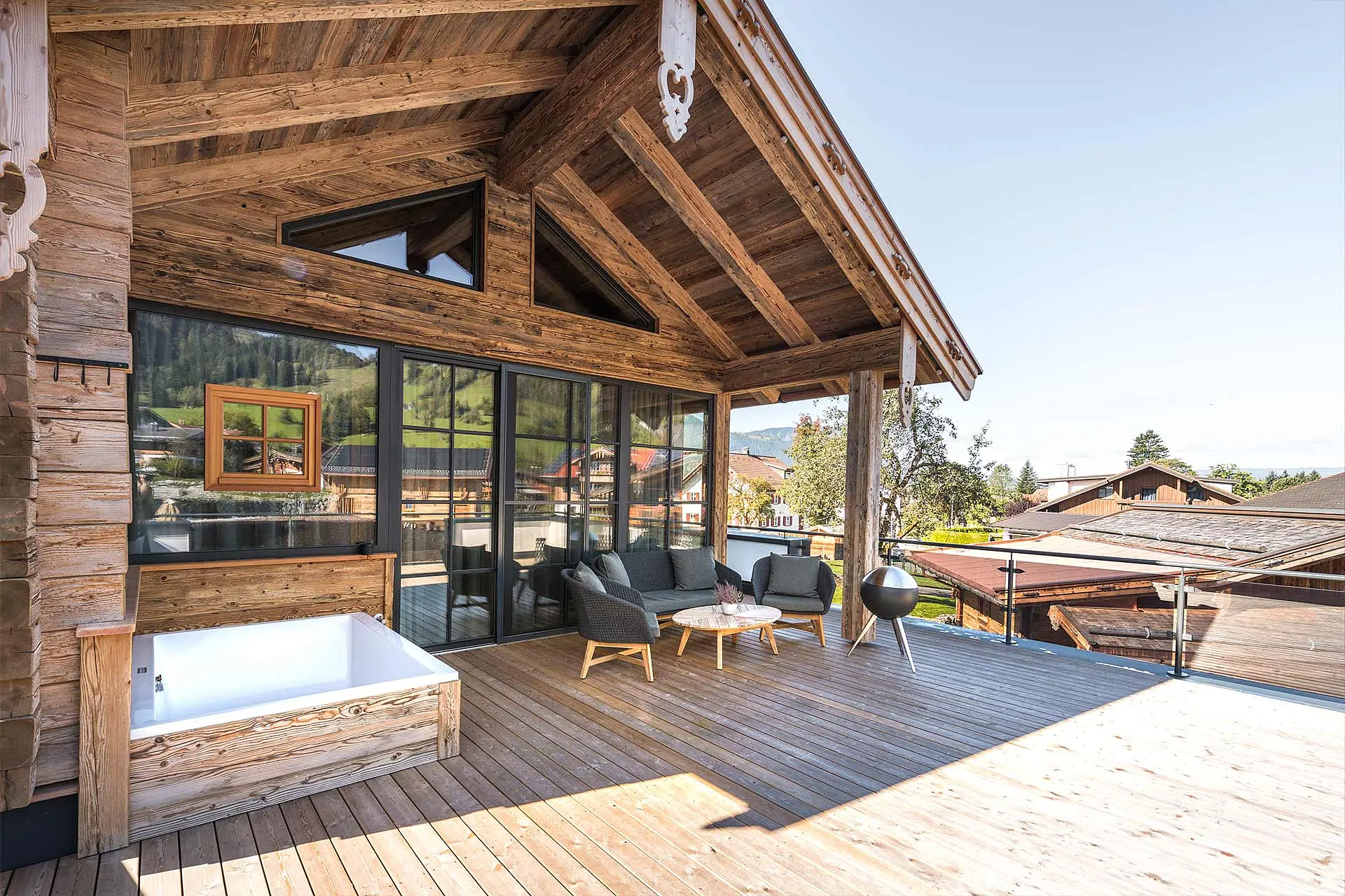 Die Terrasse eines modernen Holzhauses mit einem Dach aus sichtbarem Holz. Es gibt einen kleinen Whirlpool, gemütliche Sitzgelegenheiten und einen Außentisch. Die große Fensterfront bietet Blick auf eine Landschaft mit Bergen und Bäumen. Der Himmel ist klar und sonnig.