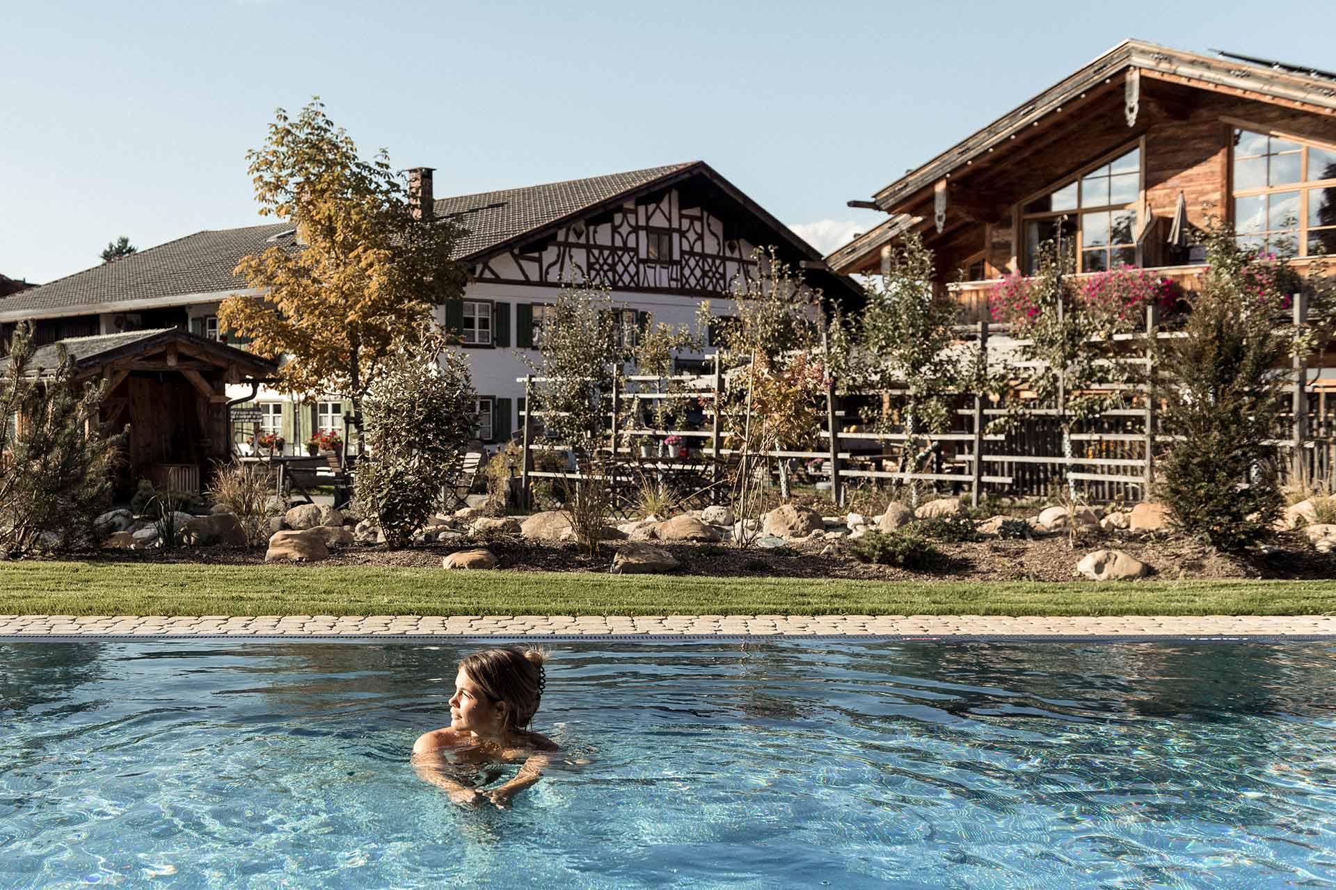 Eine Person schwimmt im klaren Wasser eines Schwimmbades. Im Hintergrund sind eine moderne Chalet-artige Unterkunft und bepflanzte Gärten zu sehen, die eine entspannte Atmosphäre vermitteln. Der Himmel ist klar und sonnig, was die idyllische Umgebung unterstreicht.