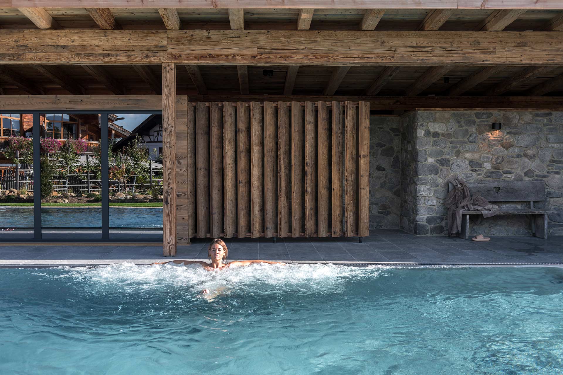 Eine Person schwimmt in einem ruhigen, klaren Pool, umgeben von einer holzverkleideten Wand und einem Steingebäude. Im Hintergrund ist eine Gartenlandschaft sichtbar. Das Bild vermittelt ein Gefühl von Entspannung und Naturverbundenheit.