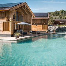 Ein modernes, aus Holz gebautes Ferienhaus liegt direkt am Rand eines klaren Schwimmbeckens. Im Vordergrund ist das Wasser sichtbar, während im Hintergrund sanfte Hügel und grüne Landschaften zu sehen sind. Sonnenschirme und Poolmöbel ergänzen die entspannende Atmosphäre.
