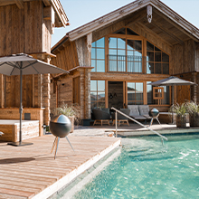 Eine moderne Holzferienunterkunft mit großen Fenstern, umgeben von einem Pool. Auf der Terrasse stehen Sonnenliegen und Sonnenschirme. Der integrierte Außenbereich bietet einen einladenden Blick auf das Wasser.