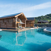 Ein modernes Holzhaus mit großen Fenstern steht in der Nähe eines klaren Schwimmbeckens. Die Umgebung ist von sanften Hügeln mit Bäumen umgeben, der Himmel ist blau und heiter. Die Architektur verbindet Natur und Komfort.