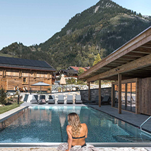 Eine Frau sitzt am Rand eines Außenpools und blickt auf eine Berglandschaft. Im Hintergrund sind Holzgebäude und Sonnenschirme sichtbar. Der Himmel ist klar, und die Szene strahlt Ruhe und Entspannung aus.