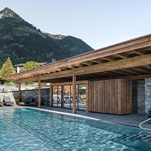 Ein modernes, einseitig offenes Gebäude aus Holz und Stein überblickt einen klaren Pool. Im Hintergrund erstrecken sich majestätische Berge, während der Außenbereich mit Liegen zur Entspannung einlädt. Die Szenerie vermittelt eine ruhige, naturverbundene Atmosphäre.