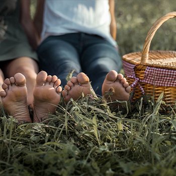 Zwei Paare nackter Füße liegen im Gras, während die Personen im Hintergrund auf einer Wiese sitzen. Neben ihnen steht ein Picknickkorb mit einem karierten Stoff. Die Szene strahlt eine entspannte und gemütliche Atmosphäre aus.