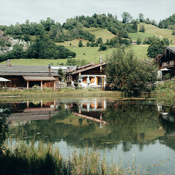 Eine idyllische Landschaft mit mehreren Holzhäusern am Ufer eines ruhigen Teichs. Im Hintergrund erheben sich grüne Hügel, die von Bäumen gesäumt sind. Die Häuser spiegeln sich sanft im Wasser, was eine friedliche Atmosphäre schafft.