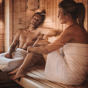 Zwei Personen entspannen in einer Sauna. Sie sitzen auf einer Holzbank, umhüllt von Handtüchern. Die Atmosphäre ist warm und entspannt, was eine ruhige und erholsame Umgebung vermittelt.