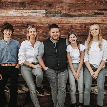 Fünf Personen stehen fröhlich vor einer rustikalen Holzwand. Sie tragen lässige Kleidung und sind gut gelaunt. Die Gruppe besteht aus drei Jugendlichen und zwei Erwachsenen, die sich nahe beieinander positioniert haben.