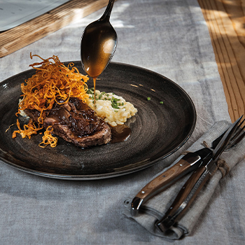 Ein Teller mit einem Stück Fleisch, garniert mit knusprigen Röstzwiebeln und einem Klecks Kartoffelpüree. Eine dunkle Sauce wird über das Fleisch gegossen. Der Teller ist auf einem Tisch mit einem Tischläufer und Besteck positioniert.